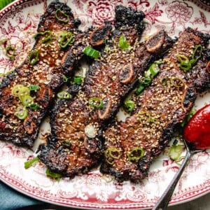 Photo shows LA galbi (korean short ribs) barbequed and served on a big white plate with side dishes on the side.