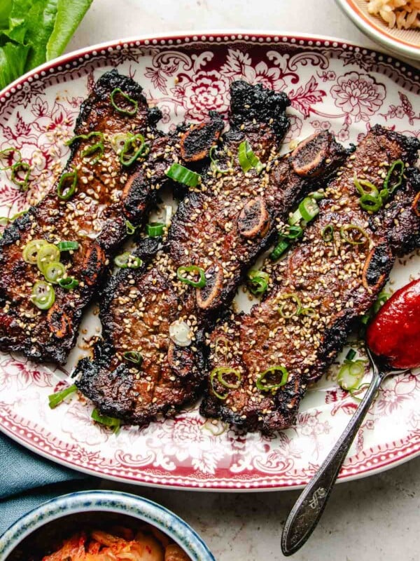 Photo shows LA galbi (korean short ribs) barbequed and served on a big white plate with side dishes on the side.