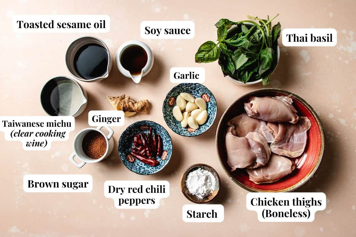 A flat lay of ingredients for Taiwanese three cup chicken in bowls and cups labeled: toasted sesame oil, soy sauce, Thai basil, garlic, boneless chicken thighs, starch, dry red chili peppers, brown sugar, ginger, and Taiwanese michiu (cooking wine).