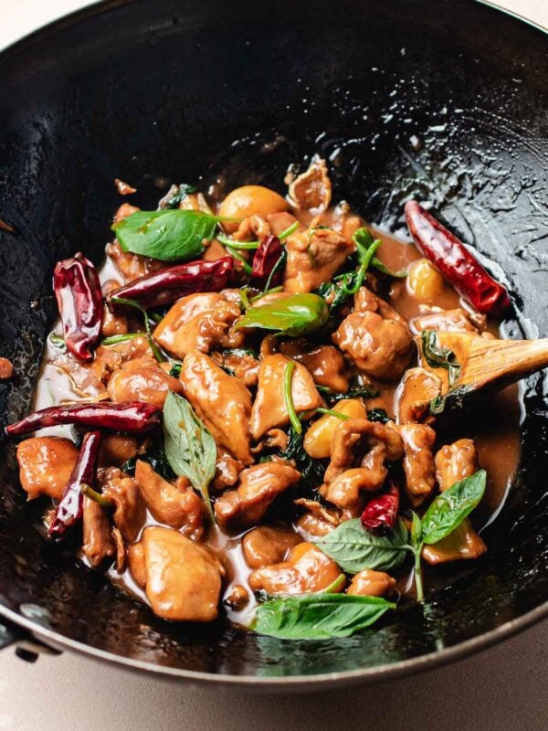 A close-up of a wok filled with Taiwanese Three Cup Chicken, dried red chilies, fresh basil leaves, and a glossy brown sauce, with a wooden spoon resting inside.