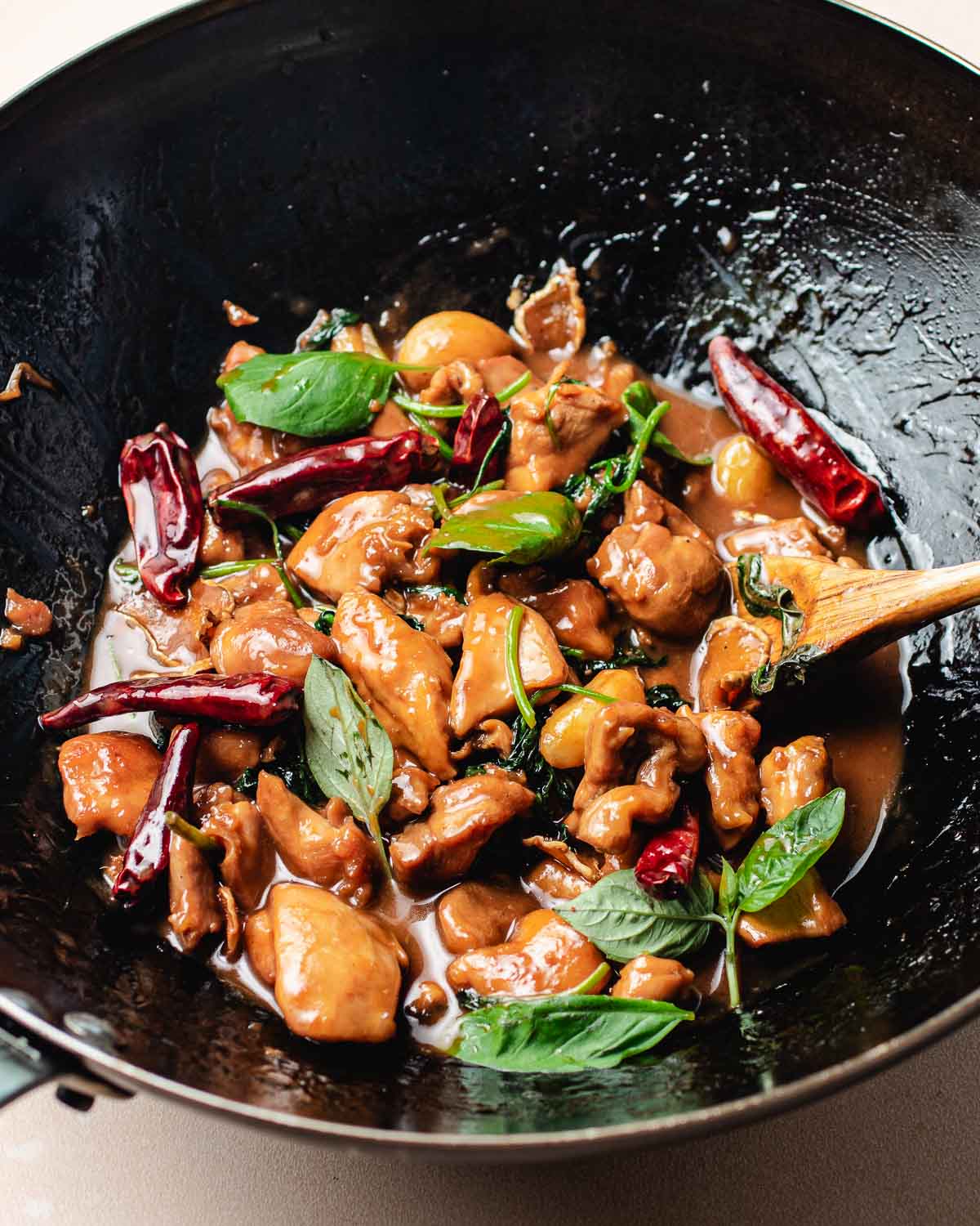 A close-up of a wok filled with Taiwanese Three Cup Chicken, dried red chilies, fresh basil leaves, and a glossy brown sauce, with a wooden spoon resting inside.