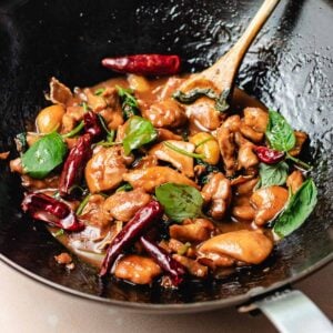 A wok filled with Taiwanese three cup chicken, stir-fried with red chili peppers, garlic cloves, fresh basil leaves, and a glossy brown sauce, with a wooden spoon resting inside.