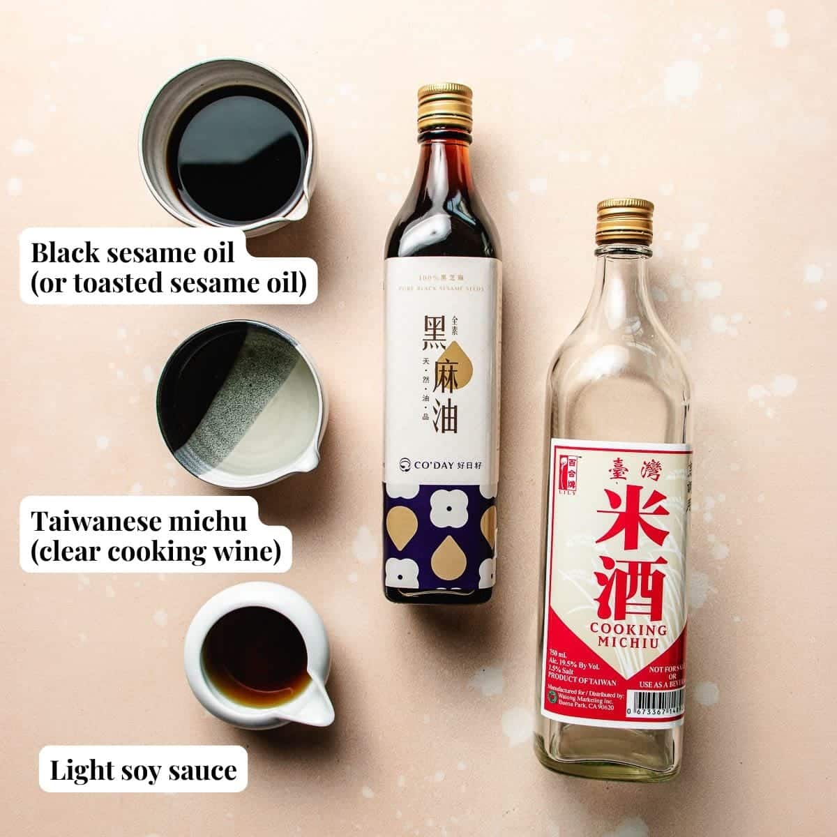 Two glass bottles—one labeled black sesame oil and the other cooking michiu—are arranged on a light surface, alongside small bowls of black sesame oil, michiu, and soy sauce. These ingredients are essential for making Taiwanese three cup chicken.