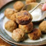 A close-up of a plate of golden-brown Paleo Meatballs, with one meatball being held up by a wooden pick. A small bowl of white dipping sauce is visible in the background.