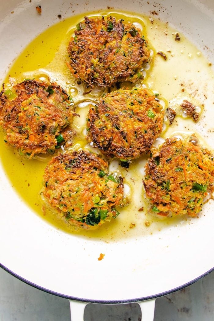 Photo shows five vegetable fritters pan fried in a large white skillet with oil bubbling