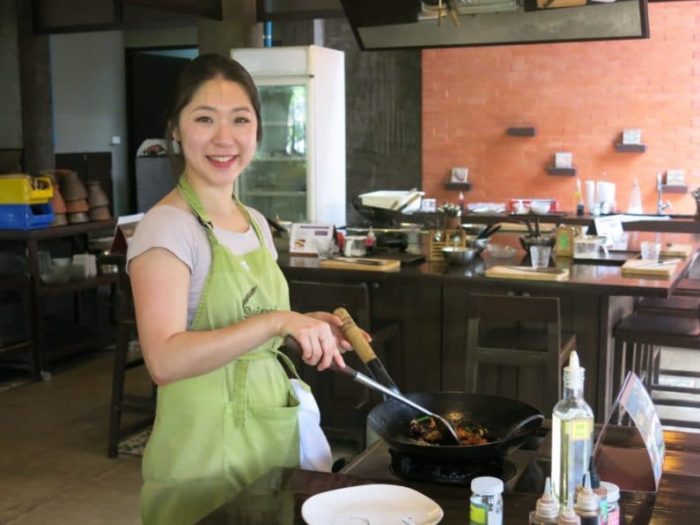 A smiling ChihY wearing a green apron stirs food in a wok at a restaurant kitchen counter.
