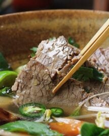 Person shows picking up a sliced brisket soaked in the pho broth with herbs in the bowl