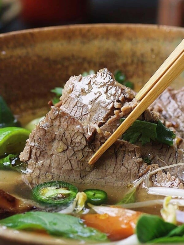 Person shows picking up a sliced brisket soaked in the pho broth with herbs in the bowl