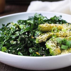 A white bowl filled with vibrant Tuscan kale salad, featuring chopped kale and sliced avocado, topped with sesame seeds and chopped green onions. The fresh salad is displayed on a wooden table.