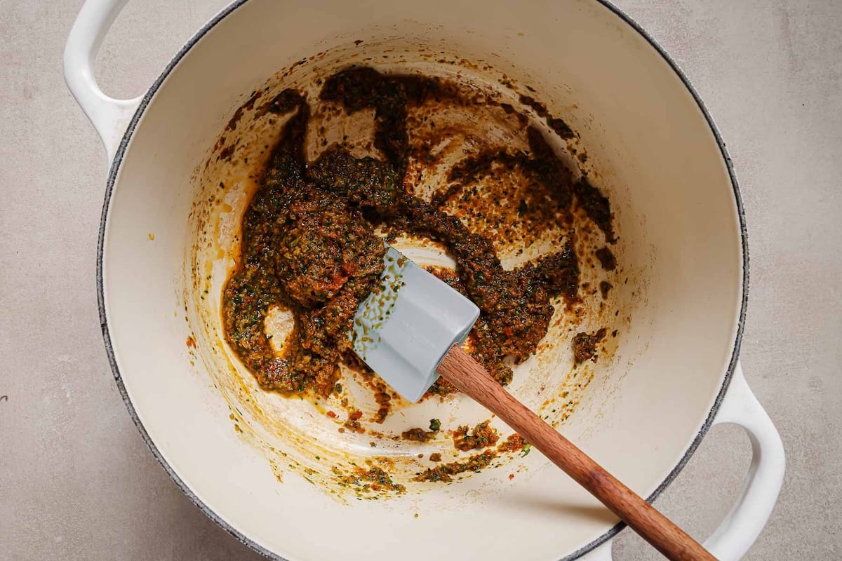A white pot with a small amount of browned herb and spice mixture stuck to the bottom, being stirred by a gray spatula with a wooden handle—perfect for making Dutch oven whole chicken.