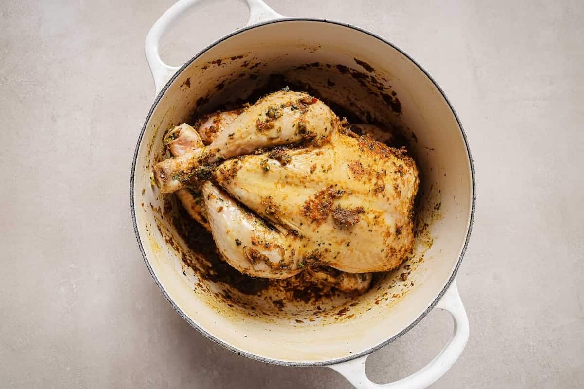 A whole chicken Dutch oven dish coated with seasoning and herbs sits in a white pot on a light-colored surface.