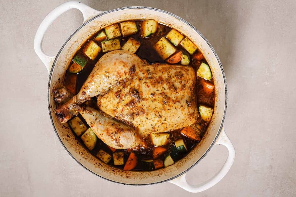 A Dutch oven whole chicken, seasoned with herbs, sits in a white pot surrounded by chunks of zucchini and carrots in a rich, dark broth. The dish rests on a light-colored surface.