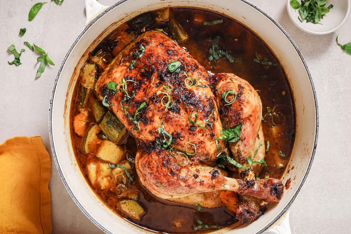 A Dutch oven roasted whole chicken sits in a white pot surrounded by vegetables and broth, garnished with fresh herbs. The chicken has golden, crispy skin, and chopped herbs are sprinkled on top, with more scattered nearby on the table.