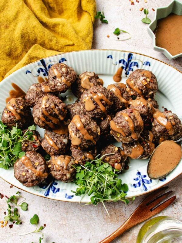 A platter of cumin lamb meatballs drizzled with peanut sauce, garnished with fresh greens on a decorative oval plate. A spoon, mustard sauce, a fork, and a yellow napkin are arranged around the dish.