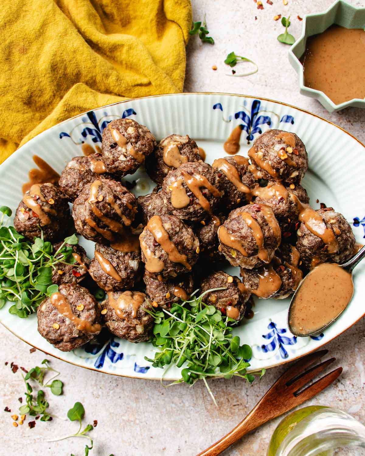 A platter of cumin lamb meatballs drizzled with peanut sauce, garnished with fresh greens on a decorative oval plate. A spoon, mustard sauce, a fork, and a yellow napkin are arranged around the dish.