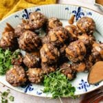 A plate of cumin lamb meatballs drizzled with peanut sauce, garnished with microgreens and red pepper flakes, with a small spoonful of extra sauce on the side—a savory nod to Xi'an flavors.