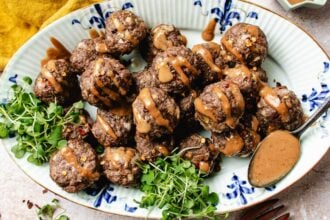 A platter of cumin lamb meatballs drizzled with Chinese peanut sauce, garnished with microgreens, and served with extra sauce on the side. The meatballs are arranged on a decorative white and blue plate.