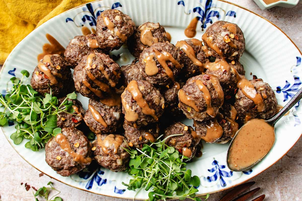 A platter of cumin lamb meatballs drizzled with Chinese peanut sauce, garnished with microgreens, and served with extra sauce on the side. The meatballs are arranged on a decorative white and blue plate.