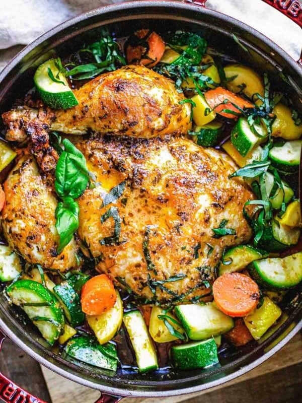 A golden brown Dutch oven whole roast chicken sits in a red pot, surrounded by cooked zucchini, yellow squash, carrots, and fresh basil leaves, with visible herbs and spices adding to its mouthwatering appeal.