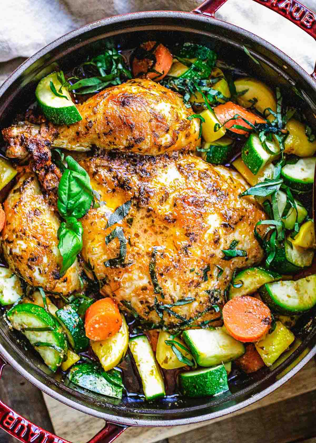A golden brown Dutch oven whole roast chicken sits in a red pot, surrounded by cooked zucchini, yellow squash, carrots, and fresh basil leaves, with visible herbs and spices adding to its mouthwatering appeal.