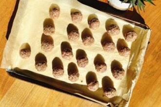 A baking sheet lined with parchment paper holds 24 evenly spaced, round cumin lamb meatballs. The tray sits on a wooden surface with sunlight casting shadows, evoking the flavors of Xi'an cuisine.