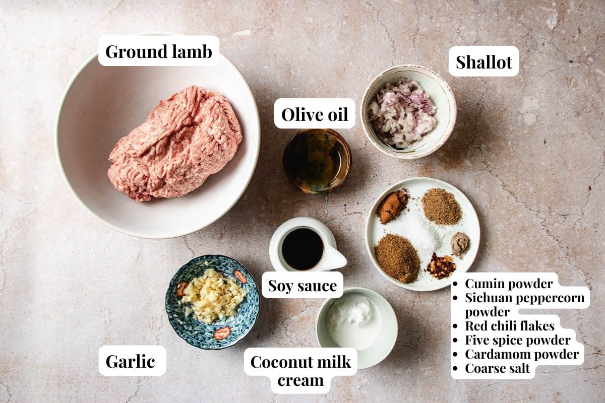 Ingredients for a recipe are arranged on a light surface, featuring ground lamb for Xi’an-style cumin lamb meatballs, chopped shallot, minced garlic, olive oil, soy sauce, coconut milk cream, and small bowls of various spices and seasonings.