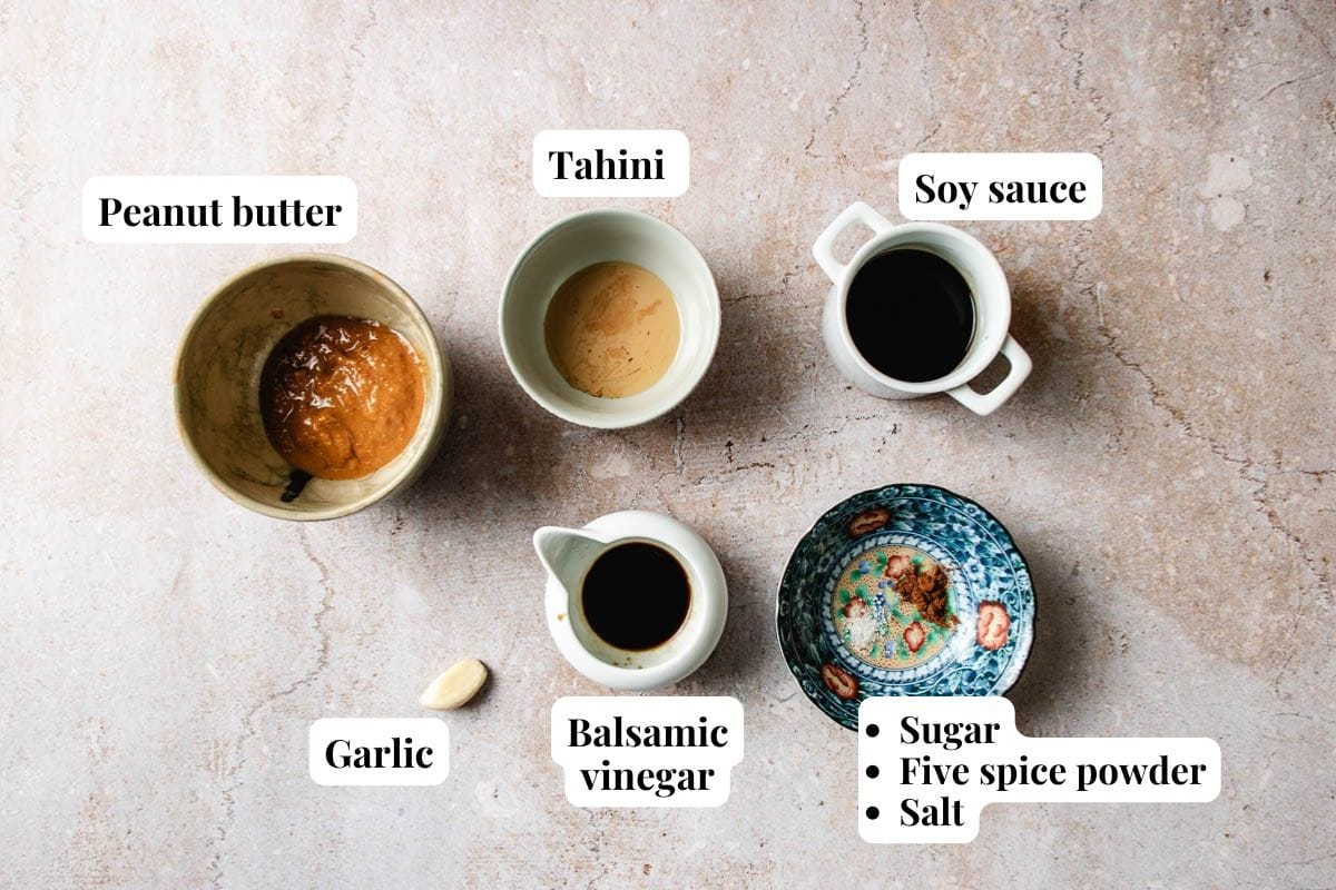 Five bowls and a garlic clove on a counter, ready for making Xi'an-inspired cumin lamb meatballs—each labeled: peanut butter, tahini, soy sauce, garlic, balsamic vinegar, and a bowl with sugar, five spice powder, and salt.