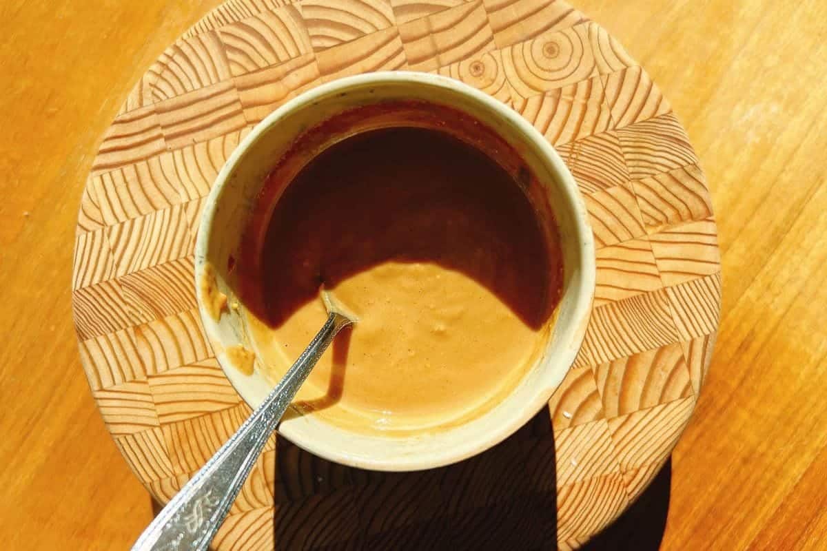 A small bowl of creamy peanut sauce with a spoon inside, placed on a round wooden board with a geometric pattern, all set on a sunlit wooden surface.