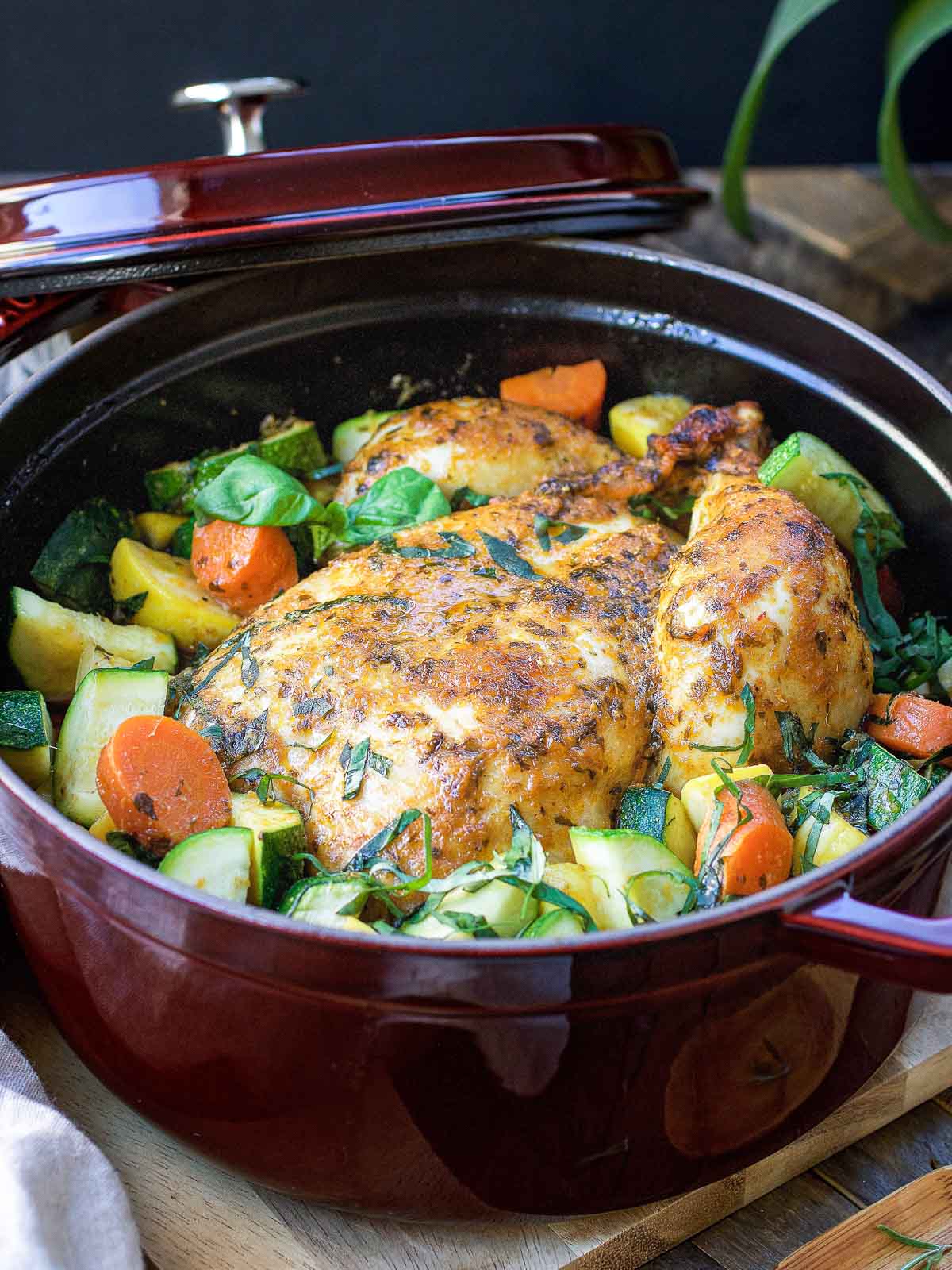 A Dutch oven roasted whole chicken, seasoned with herbs, sits in a red pot, surrounded by chopped carrots, zucchini, and fresh greens. The lid is partially open, showcasing the colorful roast chicken in this inviting Dutch oven meal.