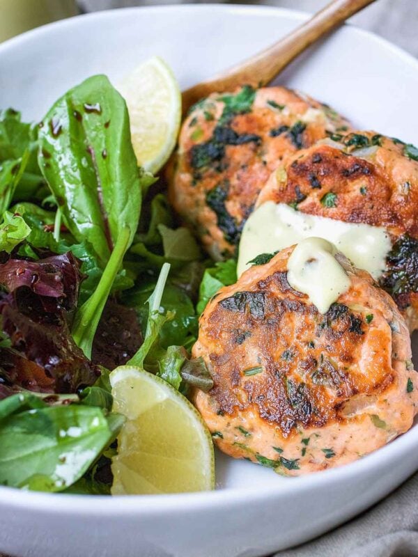 Two golden-brown Salmon Cakes with herbs, topped with a dollop of creamy sauce, served in a white bowl alongside fresh mixed greens and lime wedges—a delicious Whole30 option.