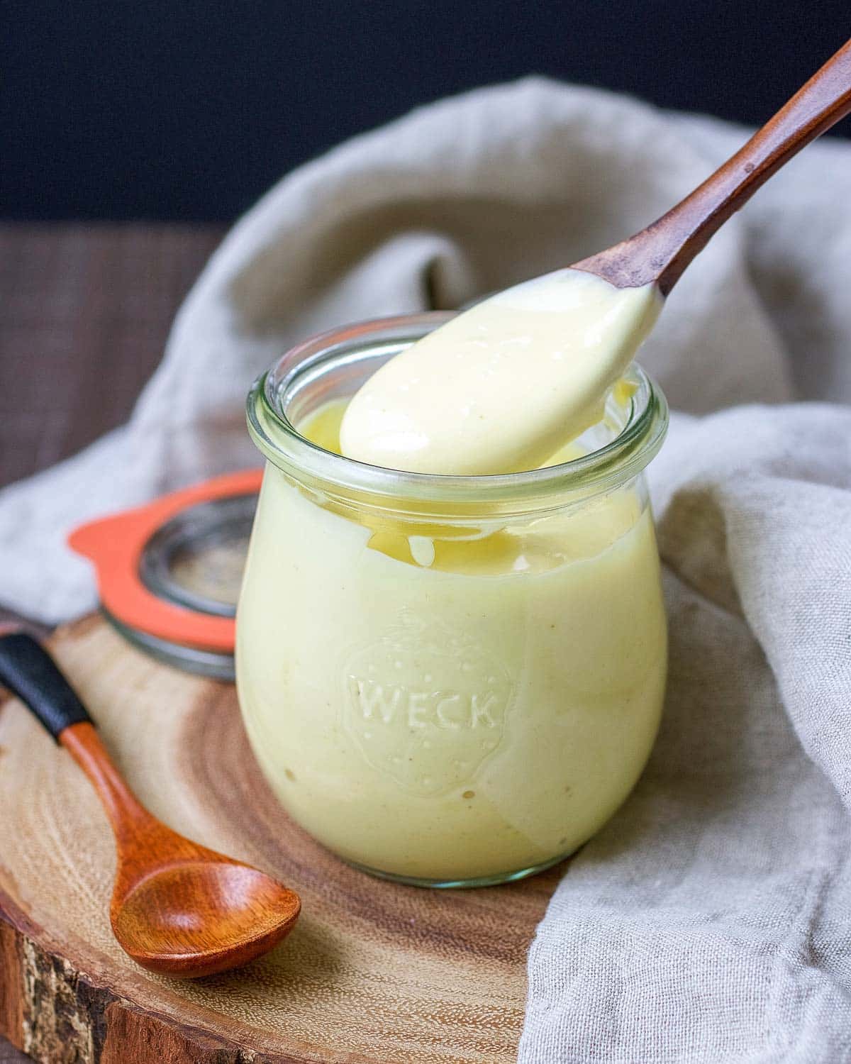 A glass jar filled with creamy Whole30 ranch dressing sits on a wooden board, with a wooden spoon lifting custard from the jar—perfect for pairing as dessert after enjoying your favorite Paleo Salmon Cakes Recipe. Another spoon and a jar lid rest nearby on a light cloth.