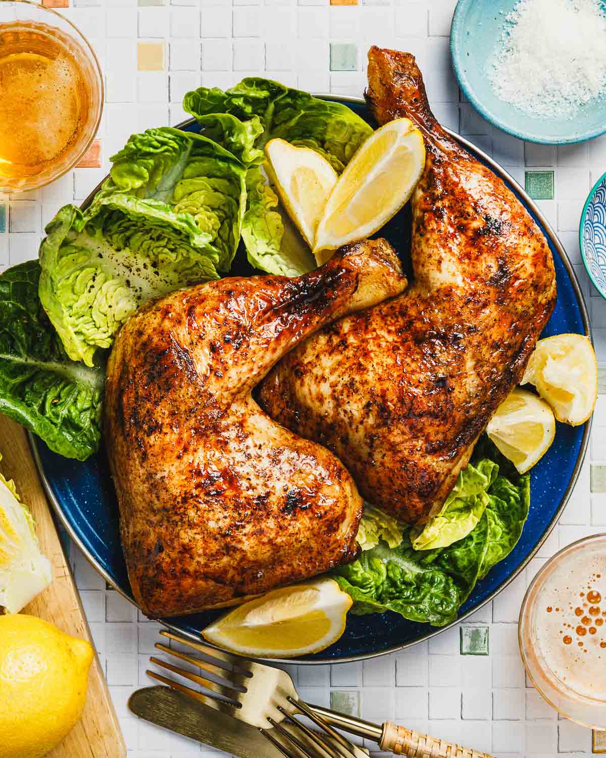 An overhead feature image shows baked lemon chicken leg quarters to golden crisp outside, served over a blue color plate with lemon and leafy greens on the side.