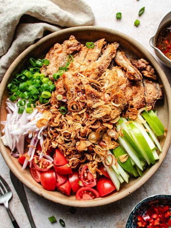 A bowl filled with Crispy Thai Chicken pieces, sliced green onions, tomatoes, cucumber, green apple, and shallots, all topped with fried shallots; a side of sweet chili dressing, a napkin, fork, and sauce bowl are placed nearby.