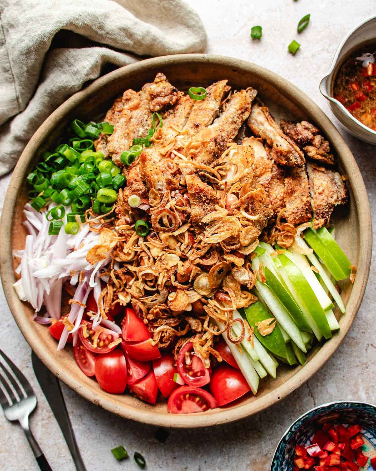 A bowl filled with Crispy Thai Chicken pieces, sliced green onions, tomatoes, cucumber, green apple, and shallots, all topped with fried shallots; a side of sweet chili dressing, a napkin, fork, and sauce bowl are placed nearby.