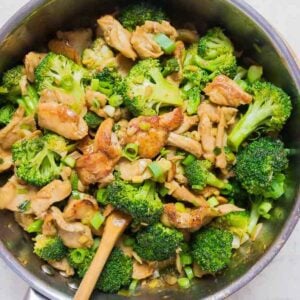 A skillet filled with stir fried tender chicken breasts, bright green broccoli florets, and chopped green onions, all mixed together with a wooden spoon for a classic Chicken and broccoli Chinese dish.