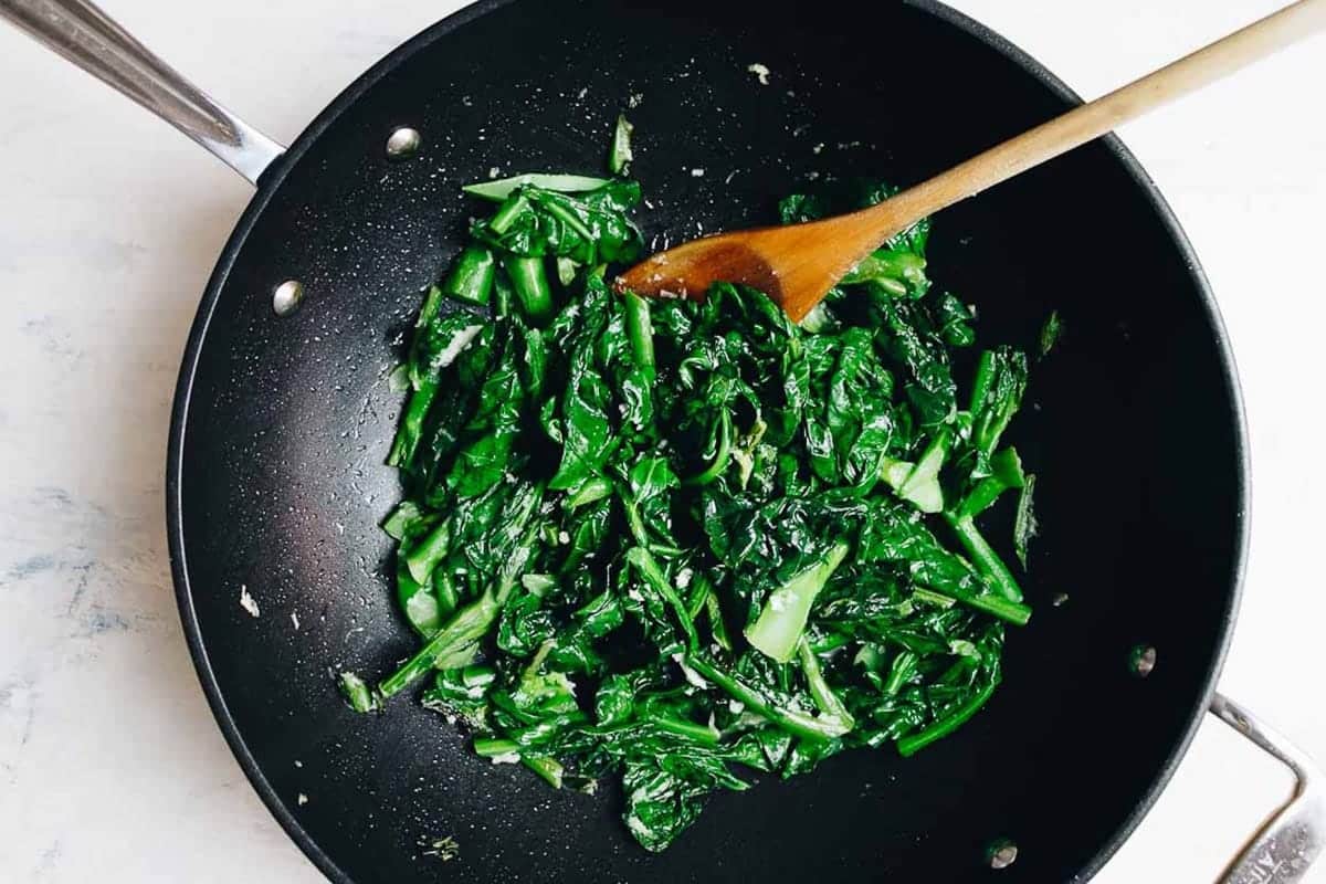 A black wok with cooked Chinese dark leafy greens - Gai Lan (Chinese Broccoli Stir Fry), and a wooden spoon, seen from above, on a light surface.