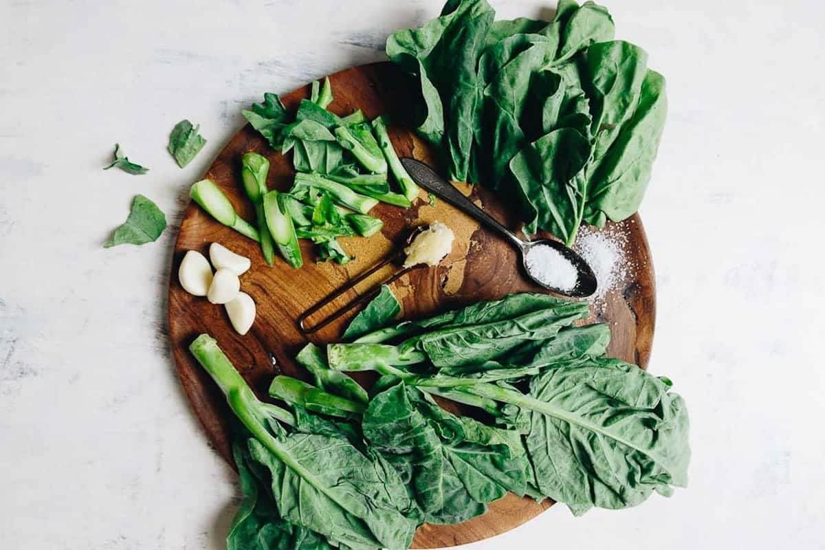 A round wooden board with fresh leafy greens—perfect for preparing Gai Lan (Chinese Broccoli)—sliced stalks, garlic cloves, ginger, and a spoon with salt on a light surface.