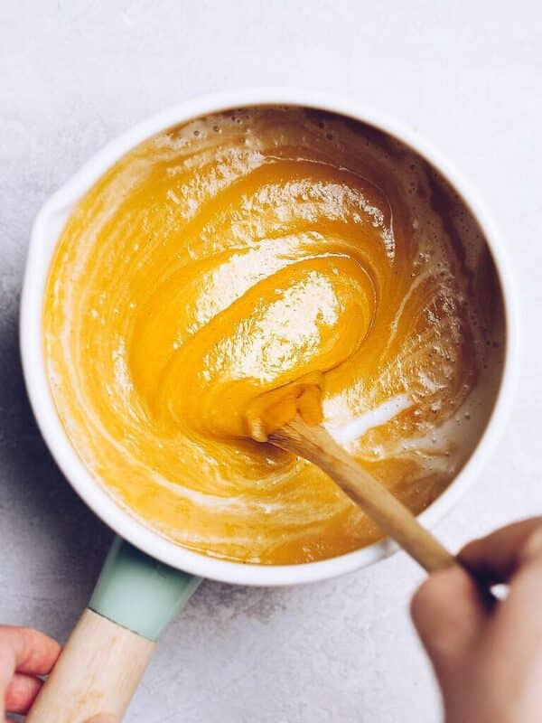 A hand stirs a thick, golden-yellow batter—reminiscent of creamy pumpkin soup—in a white mixing bowl with a wooden spoon on a light-colored surface.
