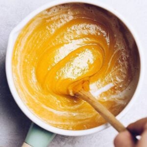 A hand stirs a thick, bright yellow quick canned pumpkin soup in a white bowl with a wooden spoon. The bowl is placed on a light surface.