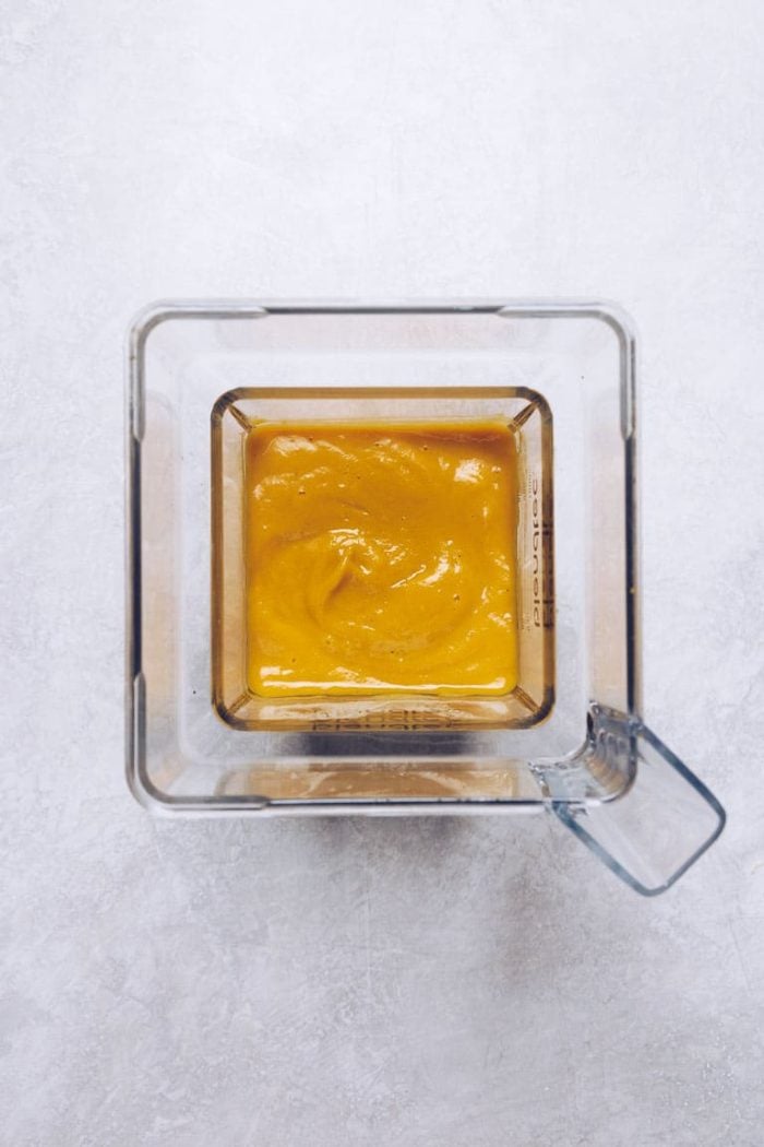 A top-down view of a blender filled with a smooth, thick yellow-pumpkin puree bisque —perfect for making quick pumpkin soup—set against a light textured background.