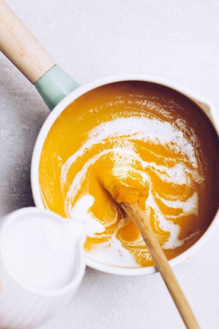 A wooden spoon stirs a pot of creamy pumpkin soup with a swirl of coconut milk cream, while someone pours more cream in from a small white pitcher, all on a light surface.