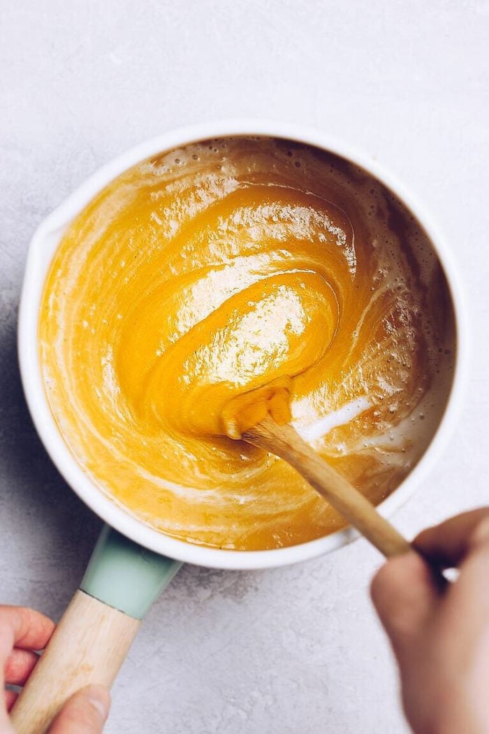 A hand stirs a thick, golden-yellow batter—reminiscent of pumpkin soup with canned pumpkin—in a white mixing bowl with a wooden spoon on a light surface.