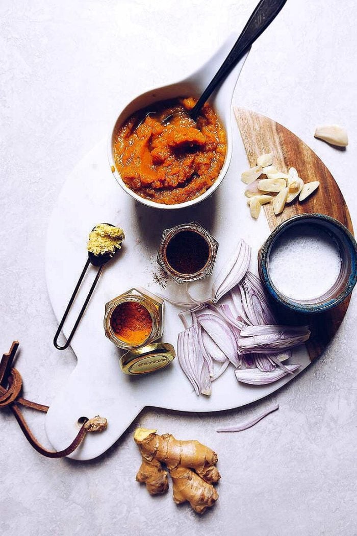 A white cutting board holds sliced onions, chopped garlic, ginger, spice jars, a bowl of curry paste with a spoon, and coconut milk—perfect essentials for making quick pumpkin soup or canned pumpkin soup at home.