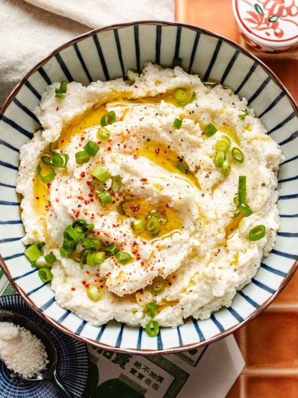A bowl of creamy cauliflower mashed potatoes, topped with chopped green onions, red pepper flakes, black pepper, and a drizzle of olive oil, served in a blue and white striped bowl on a tiled surface—a perfect low carb side.