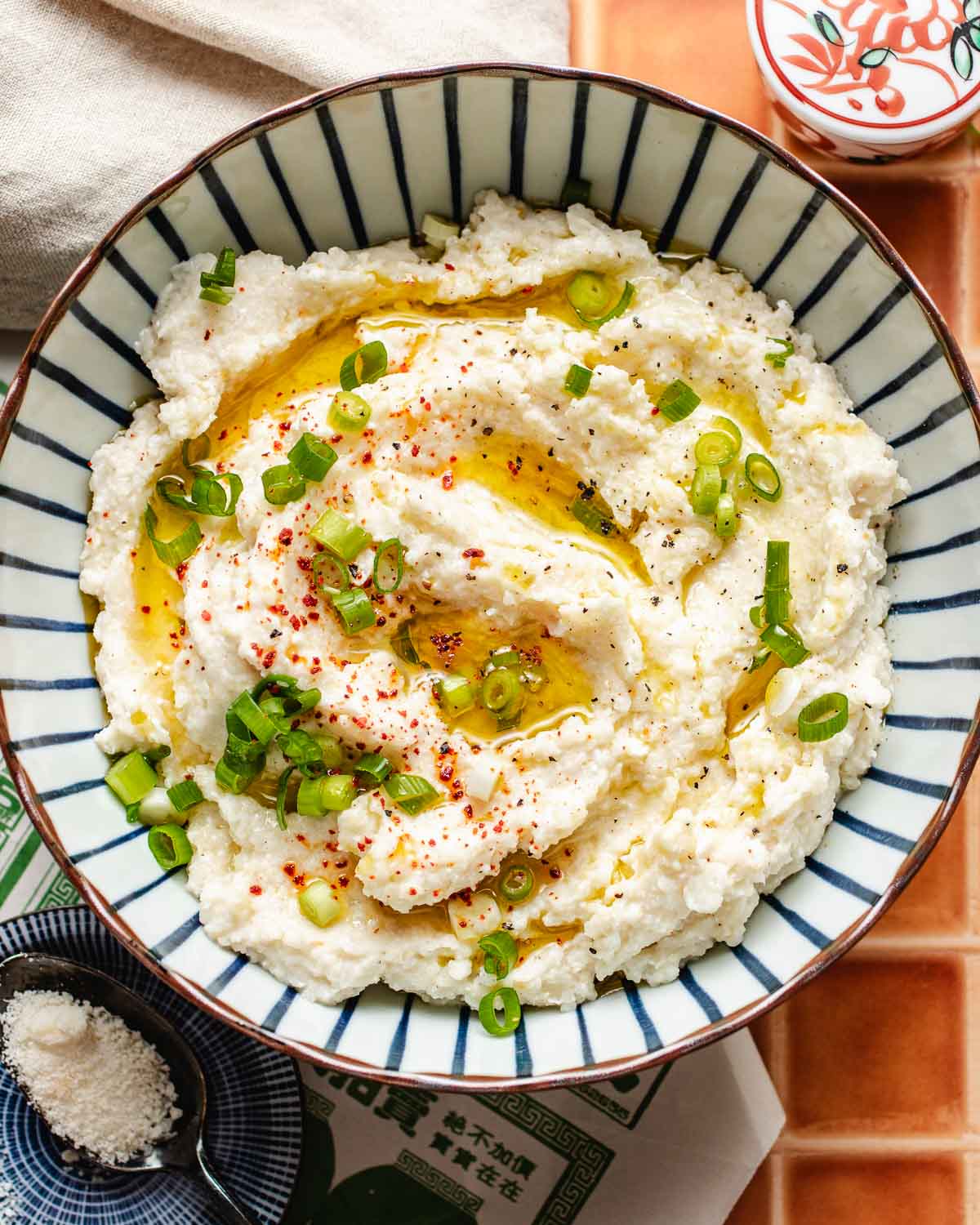 A bowl of creamy cauliflower mashed potatoes, topped with chopped green onions, red pepper flakes, black pepper, and a drizzle of olive oil, served in a blue and white striped bowl on a tiled surface—a perfect low carb side.