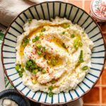 A bowl of paleo cauliflower mashed potatoes garnished with chopped green onions, red pepper flakes, black pepper, and a drizzle of olive oil, served in a striped bowl on a tiled surface.