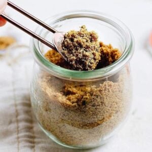 A glass jar filled with bouillon powder with a metal measuring spoon scooping some out, sits on a light-colored surface—perfect for adding to homemade chicken bouillon powder recipes.