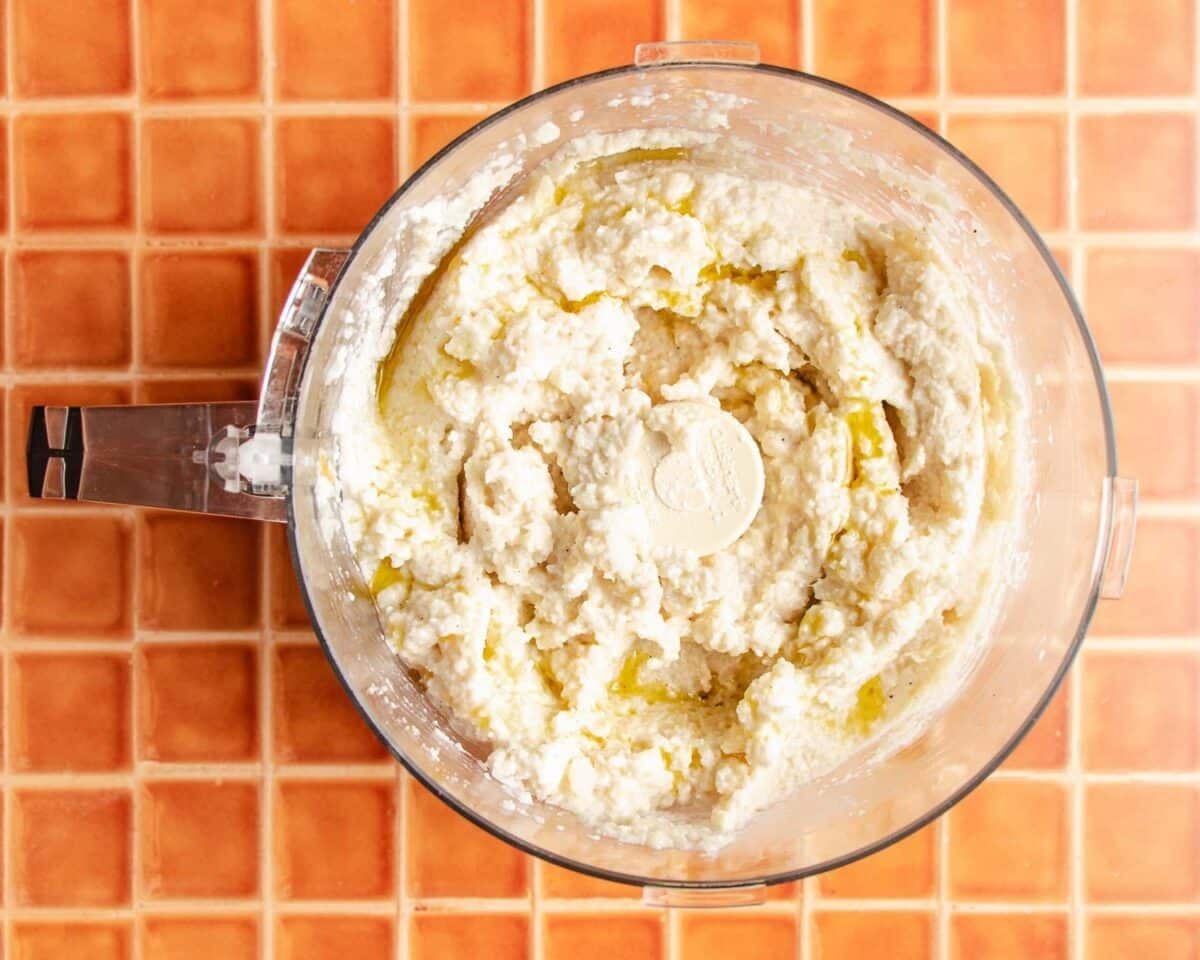 A food processor filled with a creamy, cauliflower mash with ricotta cheese mixture sits on an orange tiled countertop, photographed from above.