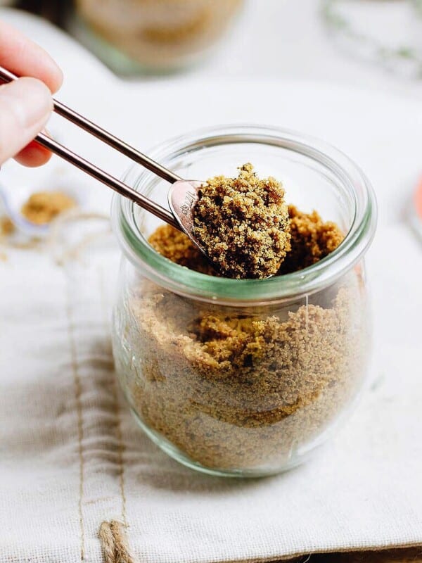 A hand holds a spoonful of homemade chicken bouillon powder above a glass jar filled with more of the savory mix, set on a light cloth.