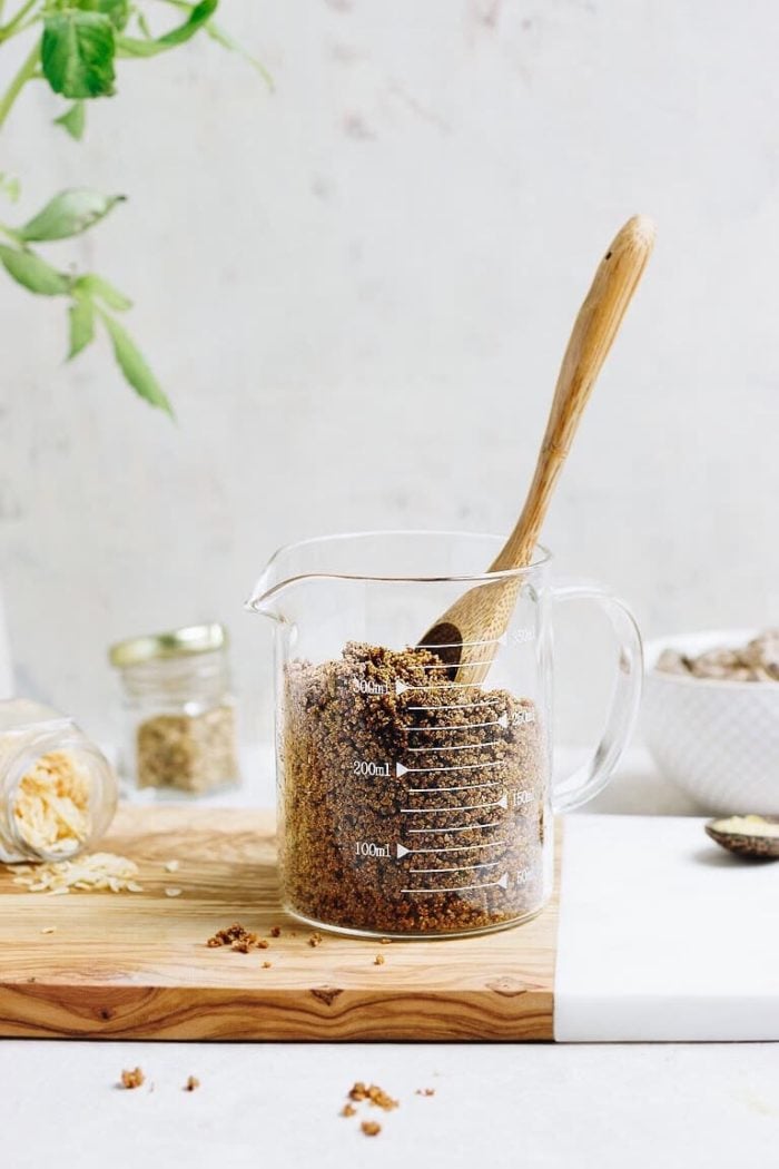 A glass measuring cup filled with bouillon powder sits on a wooden board, with a wooden spoon inside. Nearby are a jar with scattered bouillon seasoning and another small jar of homemade chicken bouillon powder, all on a light background.